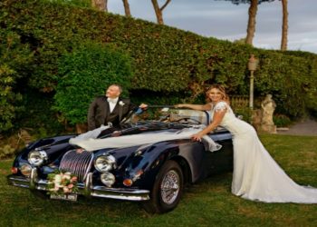 The bride and groom stand proudly next to their classic Jaguar XK150 wedding car, symbolizing love and luxury on their wedding day.