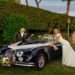 The bride and groom stand proudly next to their classic Jaguar XK150 wedding car, symbolizing love and luxury on their wedding day.