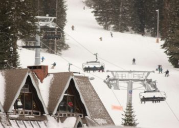 Ski lift elevated above the ground at a ski resort, with a backdrop of snowy peaks and clear blue skies.