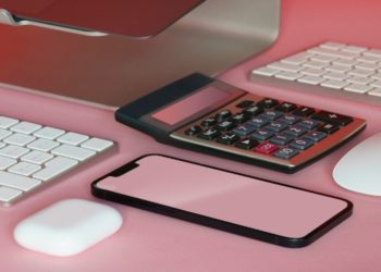 Computer monitor, calculator, keyboard, mouse, smartphone, and wireless earbuds on a pink surface, creating a modern and tech-focused scene.