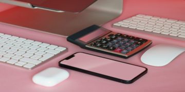 Computer monitor, calculator, keyboard, mouse, smartphone, and wireless earbuds on a pink surface, creating a modern and tech-focused scene.