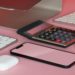 Computer monitor, calculator, keyboard, mouse, smartphone, and wireless earbuds on a pink surface, creating a modern and tech-focused scene.