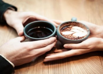 Two hands hold a gray cup of black coffee, while two hands cradle a cup of cappuccino with latte art on a wooden table. The atmosphere is warm and cozy.