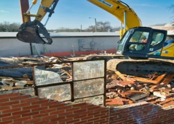Yellow excavator demolishes a brick wall at a house under demolition