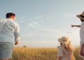 Family strolls in a vibrant field, flying a kite high in the air, with smiles on their faces and a bright sky above.