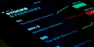 Close-up of a smartphone screen showing stock data with a dark background. Companies listed include Boeing and Disney. Graph lines suggest market trends.