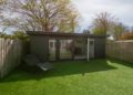 Small, modern garden room with glass doors sits in a fenced yard on green artificial grass. The scene feels peaceful and tidy under a clear sky.