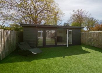 Small, modern garden room with glass doors sits in a fenced yard on green artificial grass. The scene feels peaceful and tidy under a clear sky.