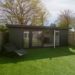 Small, modern garden room with glass doors sits in a fenced yard on green artificial grass. The scene feels peaceful and tidy under a clear sky.
