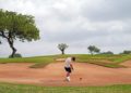 Golfer in a white shirt and shorts swings in a sandy bunker surrounded by green grass and trees under a cloudy sky, conveying focus and challenge.