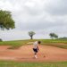 Golfer in a white shirt and shorts swings in a sandy bunker surrounded by green grass and trees under a cloudy sky, conveying focus and challenge.