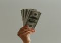 Hand is holding and fanning out several U.S. hundred-dollar bills against a plain light gray background, conveying themes of wealth and finance.