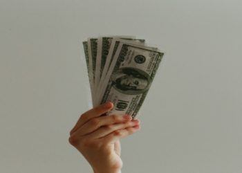 Hand is holding and fanning out several U.S. hundred-dollar bills against a plain light gray background, conveying themes of wealth and finance.