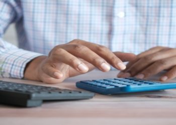 Hands of a person in a plaid shirt using a blue calculator on a wooden desk. Nearby are a keyboard, a small potted plant, and a yellow mug, suggesting a focused office setting.