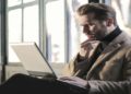 Man wearing a coat is seated on a bench, focused on his laptop in an outdoor setting.
