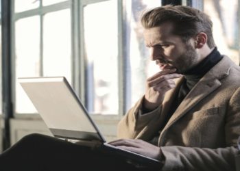 Man wearing a coat is seated on a bench, focused on his laptop in an outdoor setting.