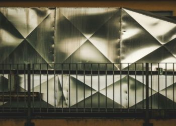 Angular, metal sheet wall with diamond patterns behind a dark railing, conveying an industrial feel. The texture is reflective and geometric.
