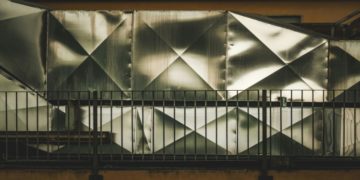 Angular, metal sheet wall with diamond patterns behind a dark railing, conveying an industrial feel. The texture is reflective and geometric.