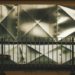 Angular, metal sheet wall with diamond patterns behind a dark railing, conveying an industrial feel. The texture is reflective and geometric.
