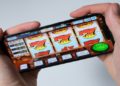Two hands hold a smartphone displaying a slot machine game with flaming symbols. A "Spin" button is visible, suggesting an engaging gaming moment.