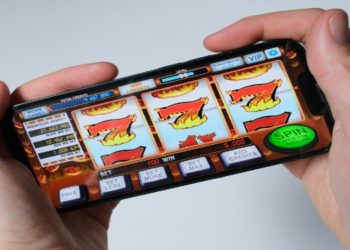 Two hands hold a smartphone displaying a slot machine game with flaming symbols. A "Spin" button is visible, suggesting an engaging gaming moment.