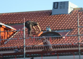 Worker repairs a red-tiled roof, balancing on scaffolding. They wear protective gear under a clear blue sky, conveying focus and diligence.