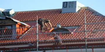Worker repairs a red-tiled roof, balancing on scaffolding. They wear protective gear under a clear blue sky, conveying focus and diligence.