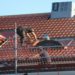 Worker repairs a red-tiled roof, balancing on scaffolding. They wear protective gear under a clear blue sky, conveying focus and diligence.