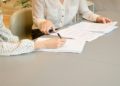 Two women at a table, engaged in signing a contract, with documents laid out in front of them.
