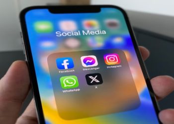 Close-up of a hand holding a smartphone showing a social media folder with app icons: Facebook, Messenger, Instagram, WhatsApp, and X. Bright, colorful screen.