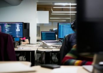 Modern office with two people working at desks, each focusing on computer screens displaying code. The room is dimly lit, creating a focused atmosphere.