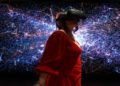 Woman in a red dress wears a VR headset, standing in front of a vibrant digital backdrop with blue and pink glowing lines, creating a futuristic feel.