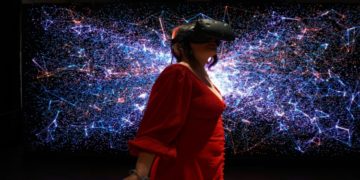 Woman in a red dress wears a VR headset, standing in front of a vibrant digital backdrop with blue and pink glowing lines, creating a futuristic feel.