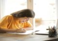 A young woman in a yellow shirt is smiling and writing notes at a wooden desk. She's focused, with a laptop and pencil holder nearby, and natural light from a window.