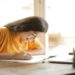 A young woman in a yellow shirt is smiling and writing notes at a wooden desk. She's focused, with a laptop and pencil holder nearby, and natural light from a window.