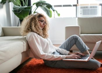 Woman seated on the floor, using on her laptop, surrounded by a comfortable and inviting space.