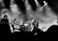 Black and white image of a rock band performing on stage. The lead singer passionately sings at the center, surrounded by dramatic smoke and dynamic lighting.