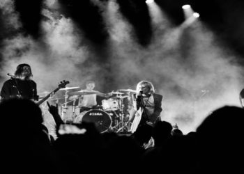 Black and white image of a rock band performing on stage. The lead singer passionately sings at the center, surrounded by dramatic smoke and dynamic lighting.