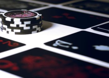 Stack of black and white poker chips sits on a table surrounded by a spread of dark playing cards adorned with red and white symbols, conveying a sense of strategy.