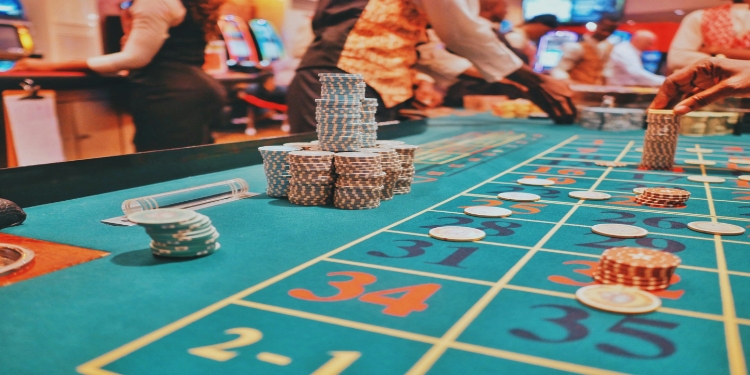 Casino table with chips stacked on a turquoise roulette mat, surrounded by people in formal attire. The scene is lively and vibrant.