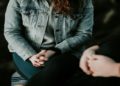 Two people sit facing each other with hands clasped. One wears a denim jacket, suggesting a serious conversation. The atmosphere feels introspective.