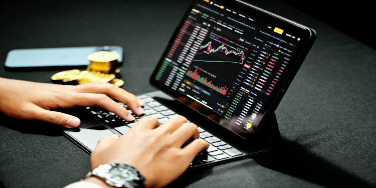 Hands typing on a laptop with a cryptocurrency trading screen. A smartphone and golden Bitcoin coins lie nearby. The mood is focused and analytical.