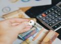 Hand holding a key over a table with Euro banknotes scattered and a calculator nearby, suggesting themes of finance, investment, or real estate.