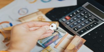 Hand holding a key over a table with Euro banknotes scattered and a calculator nearby, suggesting themes of finance, investment, or real estate.