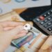 Hand holding a key over a table with Euro banknotes scattered and a calculator nearby, suggesting themes of finance, investment, or real estate.