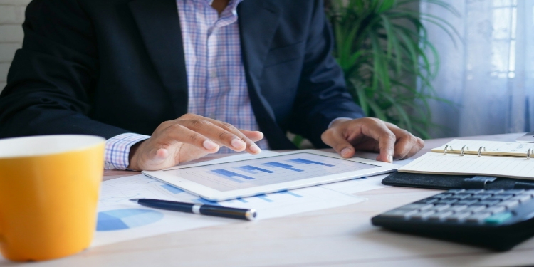 Person in a suit reviews a tablet displaying graphs at a desk, surrounded by documents, a calculator, pen, and a yellow mug. The setting is professional.
