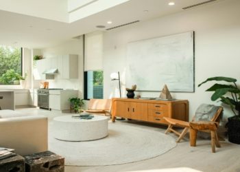 Bright, modern living room with a cream sofa, circular white rug, and a wooden sideboard. Large windows and plants add a fresh, airy feel.