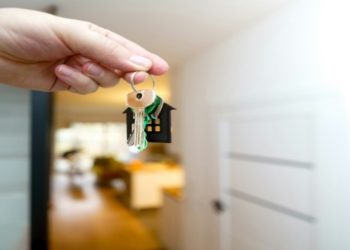 Hand holds a keychain with a house-shaped pendant and keys, symbolizing home ownership. The background shows a blurred, warmly lit interior.