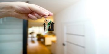 Hand holds a keychain with a house-shaped pendant and keys, symbolizing home ownership. The background shows a blurred, warmly lit interior.