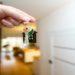Hand holds a keychain with a house-shaped pendant and keys, symbolizing home ownership. The background shows a blurred, warmly lit interior.